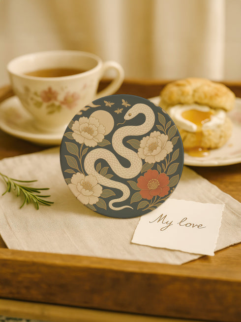 Round ceramic coaster with a chinoiserie-inspired design featuring a white snake among golden peonies, butterflies, and a glowing moon on a dark green background. Surrounded by floral and botanical accents with a soft vintage aesthetic. The coaster has a cork back and measures about 4 inches in diameter. Ideal for mugs, teacups, or wine glasses, this decorative piece suits witchy, victorian, and chinoiserie-inspired decor.